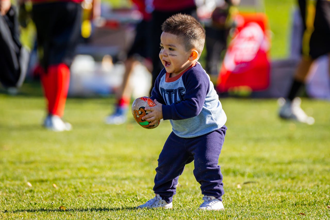 Joyful toddler playing with a football on a sunny day outdoors, dressed in casual clothes.